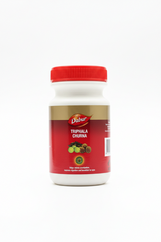 A hand holding a white plastic bottle of Dabur Triphala Churna with a red cap, set against a wooden desk background.